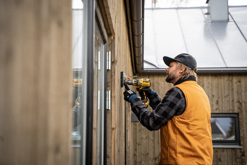 Carpenter in a work vest driving screws into vertical wood siding beside a door